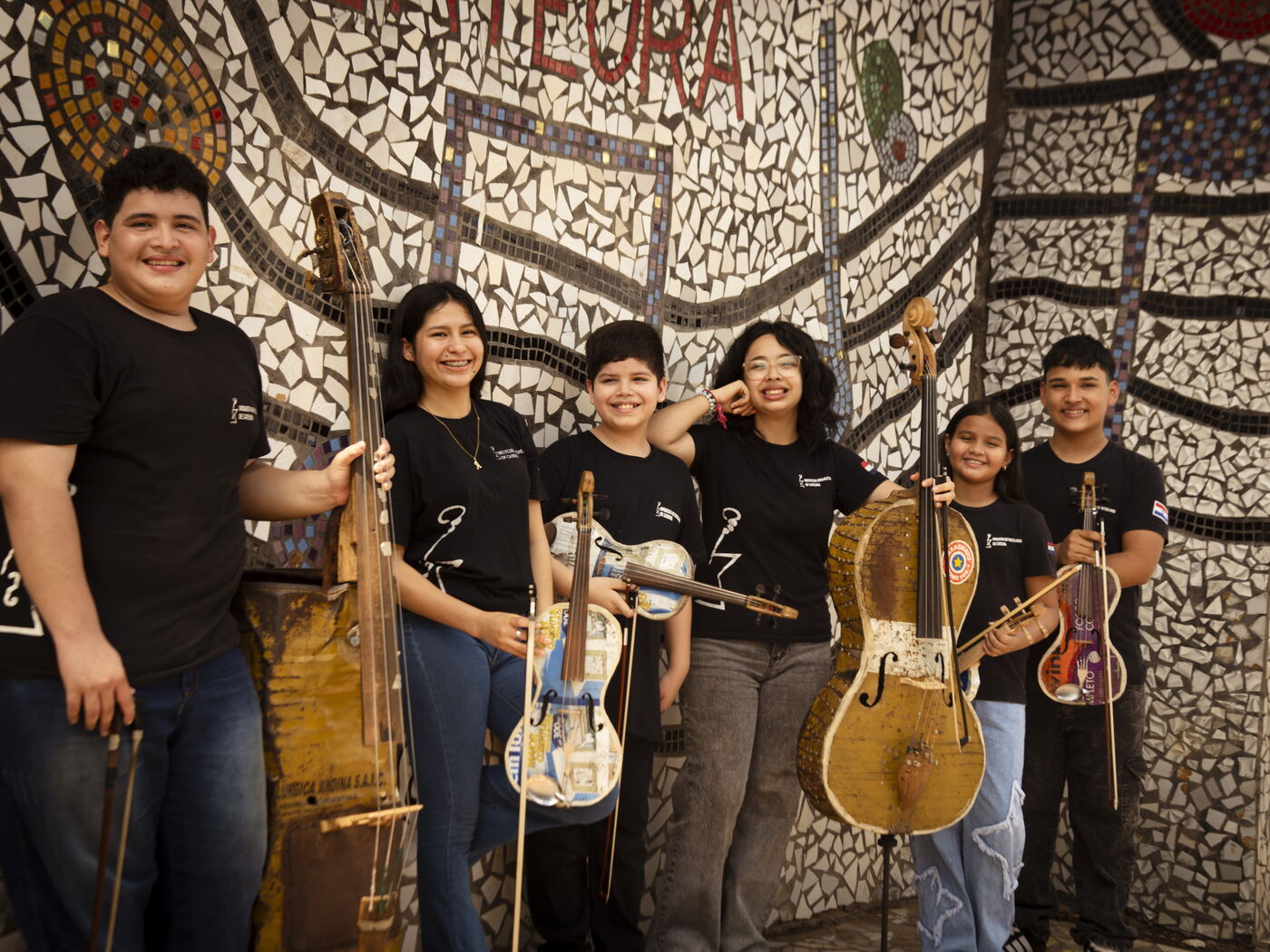 Das Orchester vor seinem Mosaik Die Protagonistinnen und Protagonisten lachen mit ihren Instrumenten vor dem Mosaik der Orquesta de Reciclados de Cateura in die Kamera